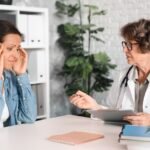 Paciente mujer madura que sufre queja de dolor de cabeza migraña menopausia, frotándose las sienes visitando a un médico, terapeuta, ginecólogo en el hospital